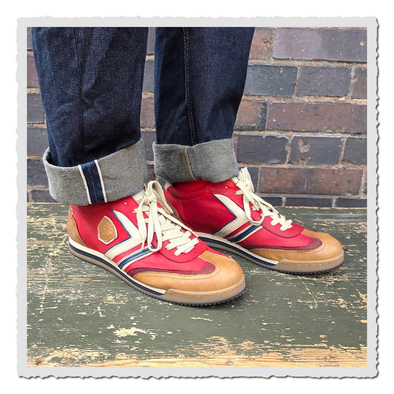 Rote und beige Sneaker an einer Person mit umgeschlagenen Jeans auf grünem Holzpodest vor Backsteinwand.